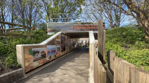 Jasmine Lowe/BBC News The viaduct has more than a dozen images on it, showing Hornsea present and past, the image has been taken on a sunny day and the leaves beside it are very green. 