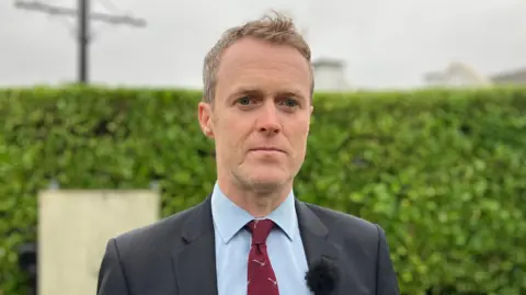 Seamus Leheny with short blonde hair is pictured staring at the camera wearing a black blazer, blue shirt and red tie.