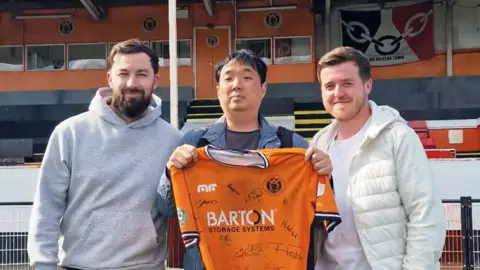 A man with dark hair and a blue jacket holding an orange football shirt between two men with brown hair and white tops with a football stand behind them