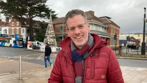 A man with dark grey hair and glasses smiles at the camera, while wearing a red puffer coat and red, blue and grey striped scarf. There is a road behind him with a tourist bus parked on it.