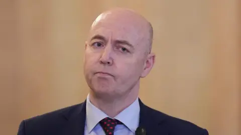PA Media Jon Burrows is bald on top with cropped hair at the side and is wearing a dark suit