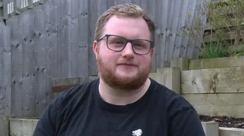 DAVE EDWARDS/BBC A man looks at the camera. He is white with brown hair and glasses. He is sitting in a garden.