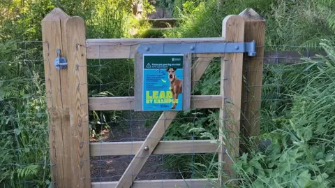 A wooden gate on a footpath surrounded by plants and hedges. The gate has a sign from the Government of Jersey on it urged dog walkers to "lead by example" and control their pets.