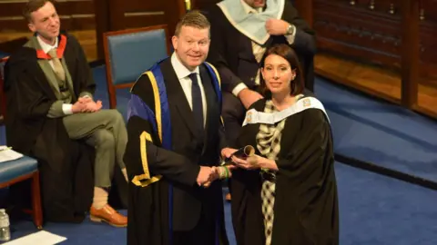 Supplied Marie Hall wearing a graduation gown and being handed a graduation scroll from a man 