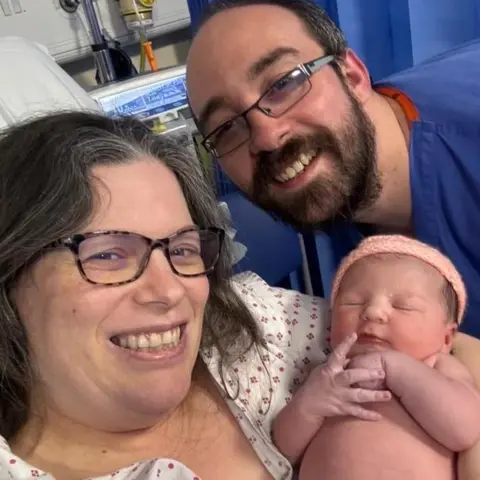 Submitted Becky Anderson and her husband Matt in hospital with their newborn. The couple smile for the camera, while the baby has her eyes closed, arms folded and wears a knitted hat 