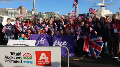 Farnham Youth Choir Members of the Farnham Youth Orchestra pose for a group photograph adorned in, and waving, Union Jack flags. They pose next to a white sign reading "Great Britain" and hold a purple banner with the 'FYC' logo on it.