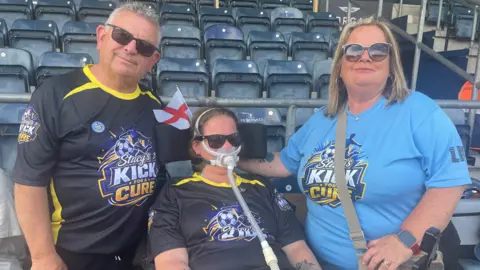 BBC Stacy Bozward is sitting down in front of rows of blue seats at a stadium. Her father, with grey and white hair, is on the left of the photo and her mother, with brown/blonde hair, is on the right. They are wearing T-shirts that include the words "Stacy's Kick for a Cure".