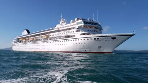 BBC White cruise ship, has many floors of windows along the side of the ship. Lifeboats can be seen attached to the side with Villa Vie Residences written in the middle and Odyssey on the front.