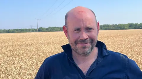 A man with his hands on his hips wearing a denim shirt and he is smiling at the camera. Crops are behind him. 
