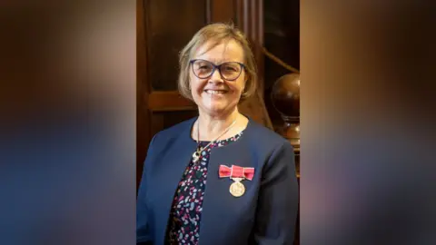Waverley Borough Council Councillor Liz Townsend, Waverley Borough Council Portfolio Holder for Planning and Economic Development, pictured receiving her British Empire Medal. Liz is wearing a navy suit jacket, patterned top and has a bob. Her British Empire Medal can be seen pinned on her jacket.