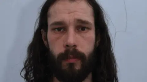 A mugshot of Robert Taylor, a bearded man with long dark brown hair