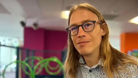 Emma Baugh/BBC John Bull wearing dark framed glasses and a blue and white floral shirt looking at camera