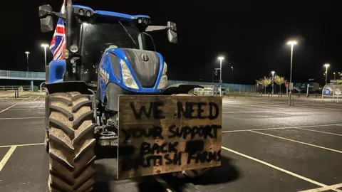 George Carden/BBC A blue tractor is parked in a supermarket car park and has a wooden sign on its hood reading: "We need your support - back British farms."