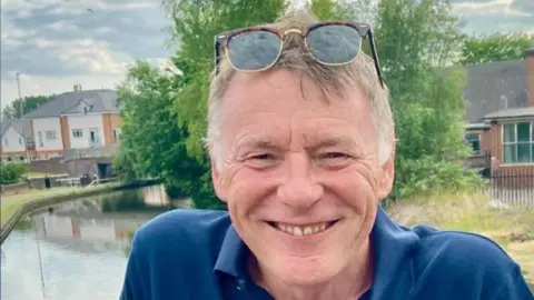 George Seward A man with grey hair and sunglasses pushed up on his head smiles at the camera. He wears a navy polo shirt. Behind him is a canal with houses and trees along the far bank.