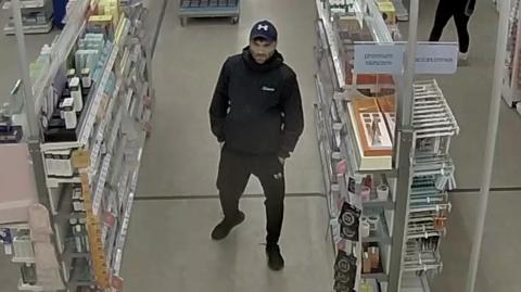 Man in a black tracksuit, with a dark Under Armour cap and black trainers. He has a short beard and moustache and his hands in his pockets. He is walking down a shop aisle.