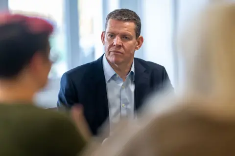 Matthew Horwood Plaid Cymru leader Rhun ap Iorwerth in conversation with an unknown person, who is out of focus in the foreground of the picture. He is wearing a dark blue suit with a light blue shirt with the top button undone and no tie