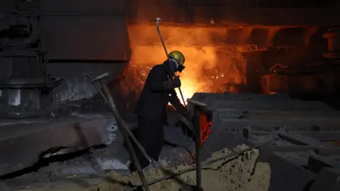 A steel worker at one of the blast furnaces at the British Steel site in Scunthorpe. The worker is wearing dark overalls, goggles and a yellow hard hat. He is standing close to a furnace which is glowing orange and emitting sparks.