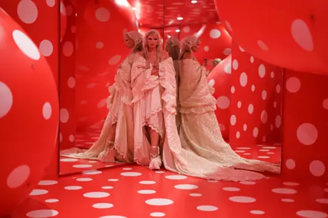 Getty Images Julia Fox in an elaborate white dress looking at the camera and reflected in mirrors in a room with red walls and giant round sculptures covered in white spots