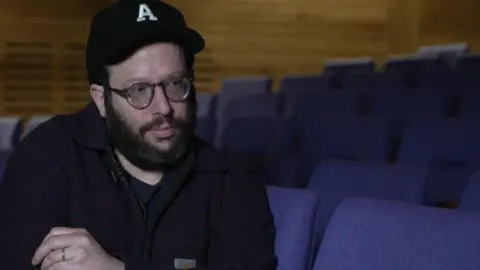 Adam Lenson sitting in theatre wearing a blue cap and glasses.
