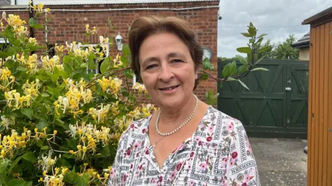 A woman with brown hair and wearing a pearl necklace and a white top with patterns on it standing in front of a bush. In the background is a gate and the back of a house.
