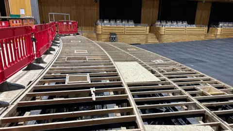 The Glasshouse The Sage One concert hall during refurbishment work. Wood floor panelling on a raised section of stage has been removed to show the supporting battens and a glimpse of the floor and supports underneath. To one side are red plastic barriers and beyond are the wood panelled side boxes where members of the audience can sit. The carpet is a dark RAF blue. 