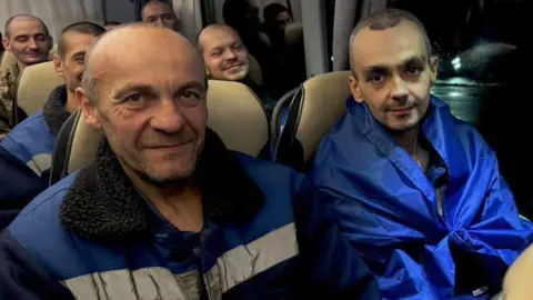 Zelenskiy/Official via Telegram Two newly released prisoners of war, dressed in blue jackets, smile at a camera while sitting on a coach 