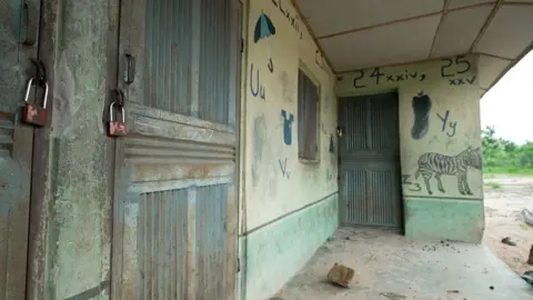 The front of a school building, the doors shut and padlocked. Children's drawings, included one of a zebra, decorate the walls