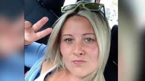 South Wales Police Joanne Penney, who has shoulder length blonde hair and is wearing a white and blue top, with yellow sunglasses on her head. She is looking at the camera and pouting, while making a peace sign with her hand.