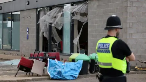 Reuters A view of damage to Holiday Inn Express hotel after rioters attacked the building in Rotherham, with chairs scattered around outside and glass from the windows smashed on to the ground. A police officer also folds his arms and looks into the distance next to the damage.