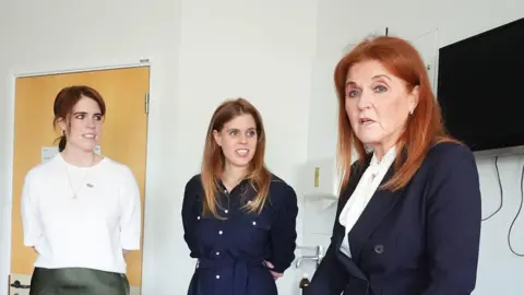 PA Media Sarah, Duchess of York and her daughters Princess Beatrice and Princess Eugenie speak to a patient  at University College Hospital, London, earlier this year.