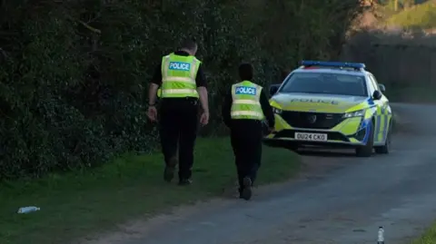 Qays Najm/BBC A yellow and blue police car is parked up on a narrow country track. Two uniformed police officers are walking towards the car with their backs turned. There is a line of trees or hedging to the left of the frame.