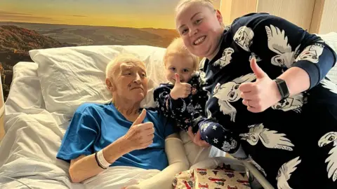 Ashgate Hospice Philip Bramall, in a hospice bed, with Esme and Faye, all giving thumbs up to the camera