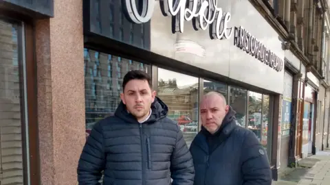 Lino and Antonio Massimo pictured outside their restaurant. Lino is tall with dark hair and dark eyes. Antonio is shorter and bald with short, grey stubble. Both men have a serious expression.