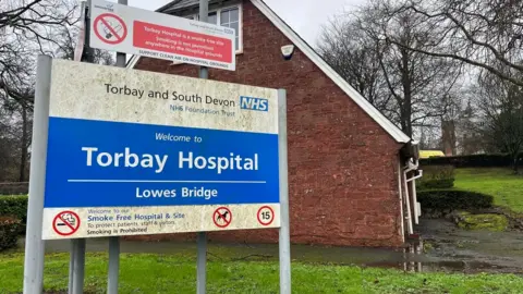 A sign for Torbay Hospital with a no-smoking sign above, in front of a house near the entrance to Torbay Hospital