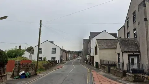 Google The A5086 Main Street in Cleator: terraced houses with slate roofs line the road on either side; there is a white car is in the distance.