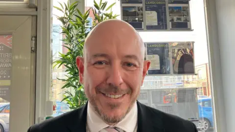 A bald, bearded man in a suit in an estate agent's office