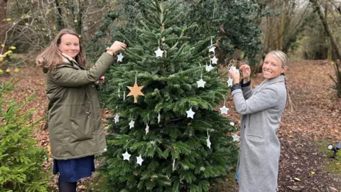 Mid and South Essex NHS Foundation Trust Two women standing in woodlands and putting Christmas decorations on a tree in the shape of white starts. They are both wearing coats and smiling at the camera.