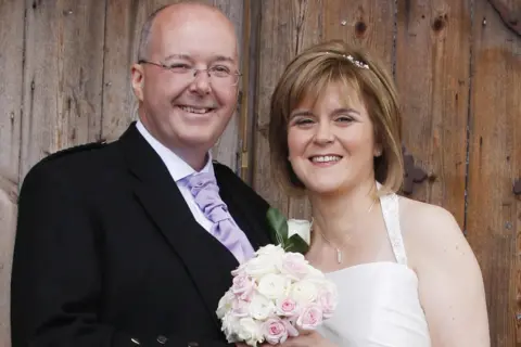 PA Media Murrell and Sturgeon in wedding attire posing in front of a wooden backdrop. He is wearing a dark suit with a light tie and buttonhole and she is in a white dress holding a bouquet of pale pink and white roses.