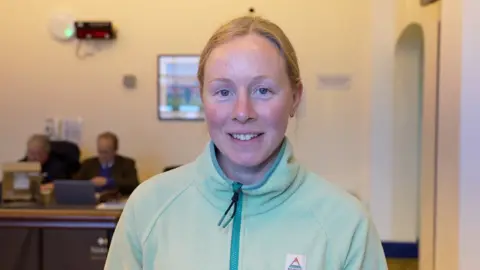 Qays Najm/BBC Katie Margarson smiles at the camera. She has blonde hair that has been tied back behind her head. She wears a light green jumper. 