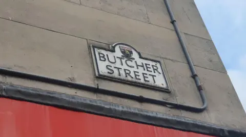 BBC News A old fashioned street sign shows the words Butcher Street. It is displayed on a grey stone building with a red base. 
