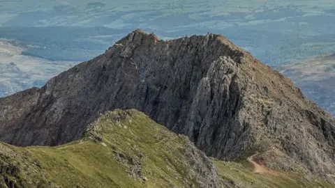 Ian Capper / Geograph A narrow and steep mountain ridge, with fields in the background. 