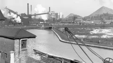A black and white photograph showing a body of water with an industrial site in the background. There are plumes of smoke or vapour emanating from the buildings. The brick building in the foreground has broken windows on its upper floor. There is also a crane in the foreground.