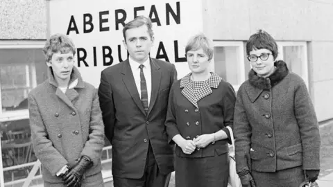 Getty Images A black and white image from the late 1960s showing four teachers standing in front of a large sign that reads 'Aberfan Tribunal'. The three female and one male teacher are in smart, formal wear. Two of the women have gloves on. Only Mair Morgan, on the right of the image, is wearing glasses. 