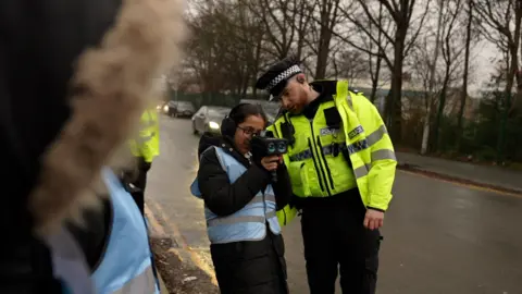A pupil wearing a high visibility jacket holds a speed camera as a police officer helps her