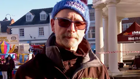 Kevin wears a blue knitted hat and brown coat with fleece inlay. He is smiling and wearing sunglasses. The sky is blue behind him.