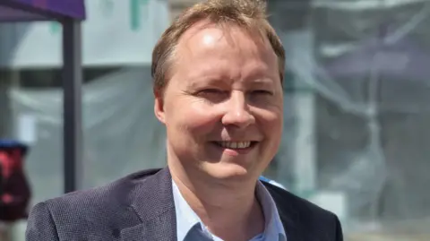 BBC A man with brown hair looking towards the camera. He's wearing a blue suite and a blue shirt. 