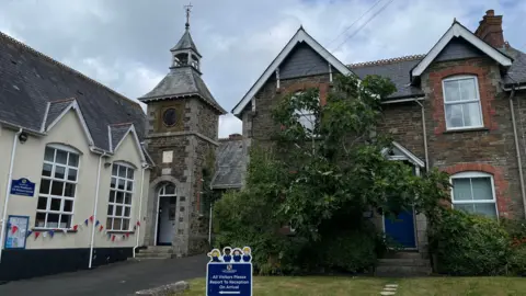  The outside of a school. The building on the left is white with large windows and bunting. The building in front is made of a brown brick and has a green bush growing on it. There are grey clouds in the sky. 
