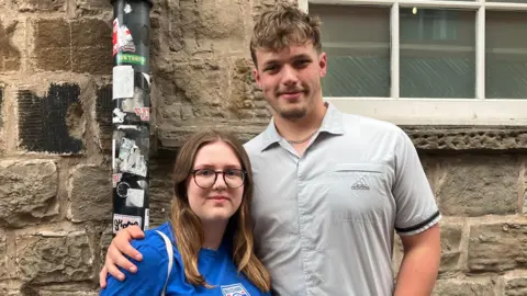 Chloe Aslett/BBC A young man with curly blond hair and short beard. He is wearing a grey button-up shirt. Next to him is Violet, about the height of his shoulders. She wears a blue t-shirt and glasses, and has long blondey-brown hair.