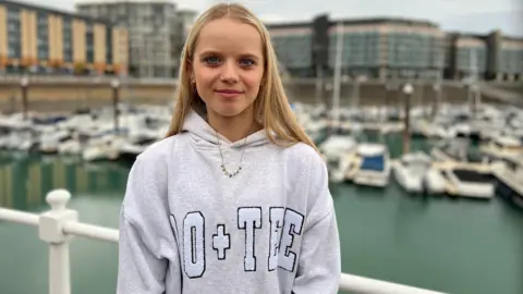 BBC Sofia smiles at the camera. She is wearing a grey hoodie with a necklace. Her blonde is beyond her shoulders. Behind her, there are boats in a marina with buildings further in the background.
