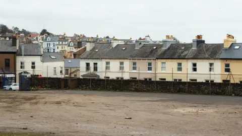Desc The site, a large empty space, with a stone wall, you can see houses in the background.